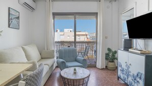 A living room with a sofa, armchair, small table, TV, and balcony with a view of buildings. - Apartment 'Playa Regla' with Balcony, Wi-Fi and Air Conditioning (Chipiona)