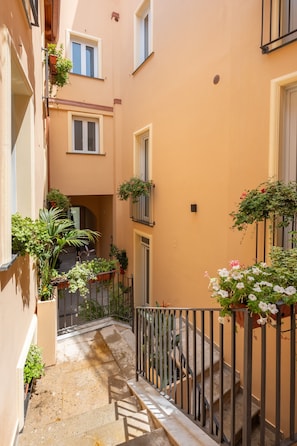 A narrow alleyway with a staircase leading to a door, surrounded by buildings with balconies adorned with plants.