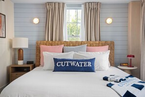 A neatly made bed with a blue pillow labeled "CUTWATER," a wooden nightstand with a lamp, and a small table with a clock. - Cutwater Inn & Suites (Newburyport)