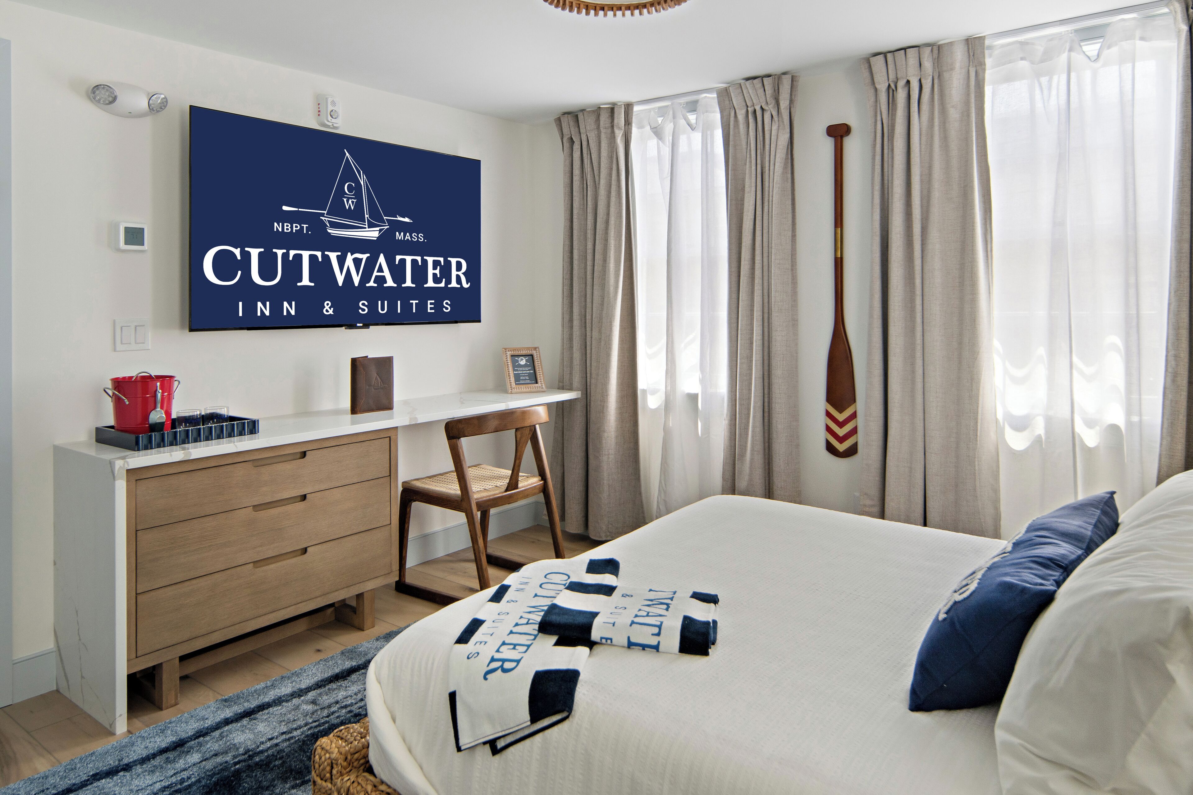 A hotel room with a bed, a desk, a chair, a television displaying the hotel name, and a window with curtains.