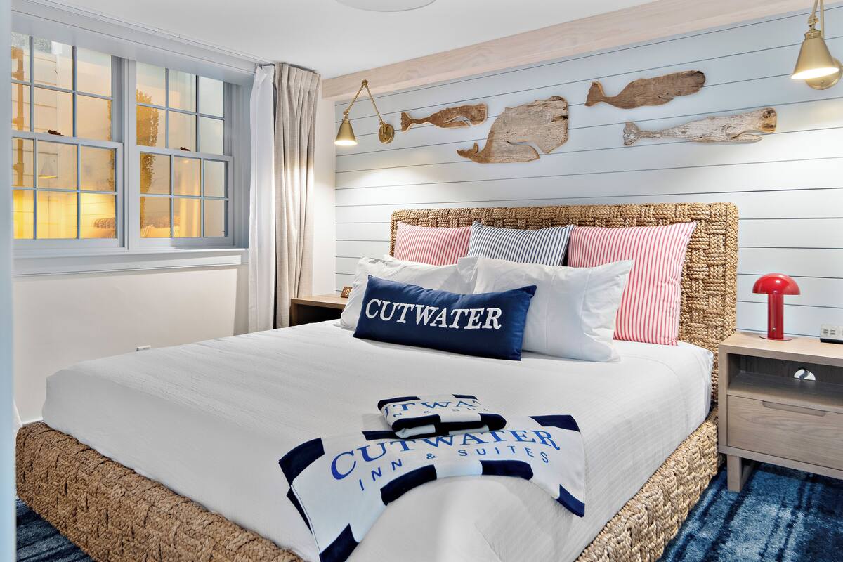 A bed with a white and blue bedspread, a headboard with a woven texture, and a nightstand with a red lamp.