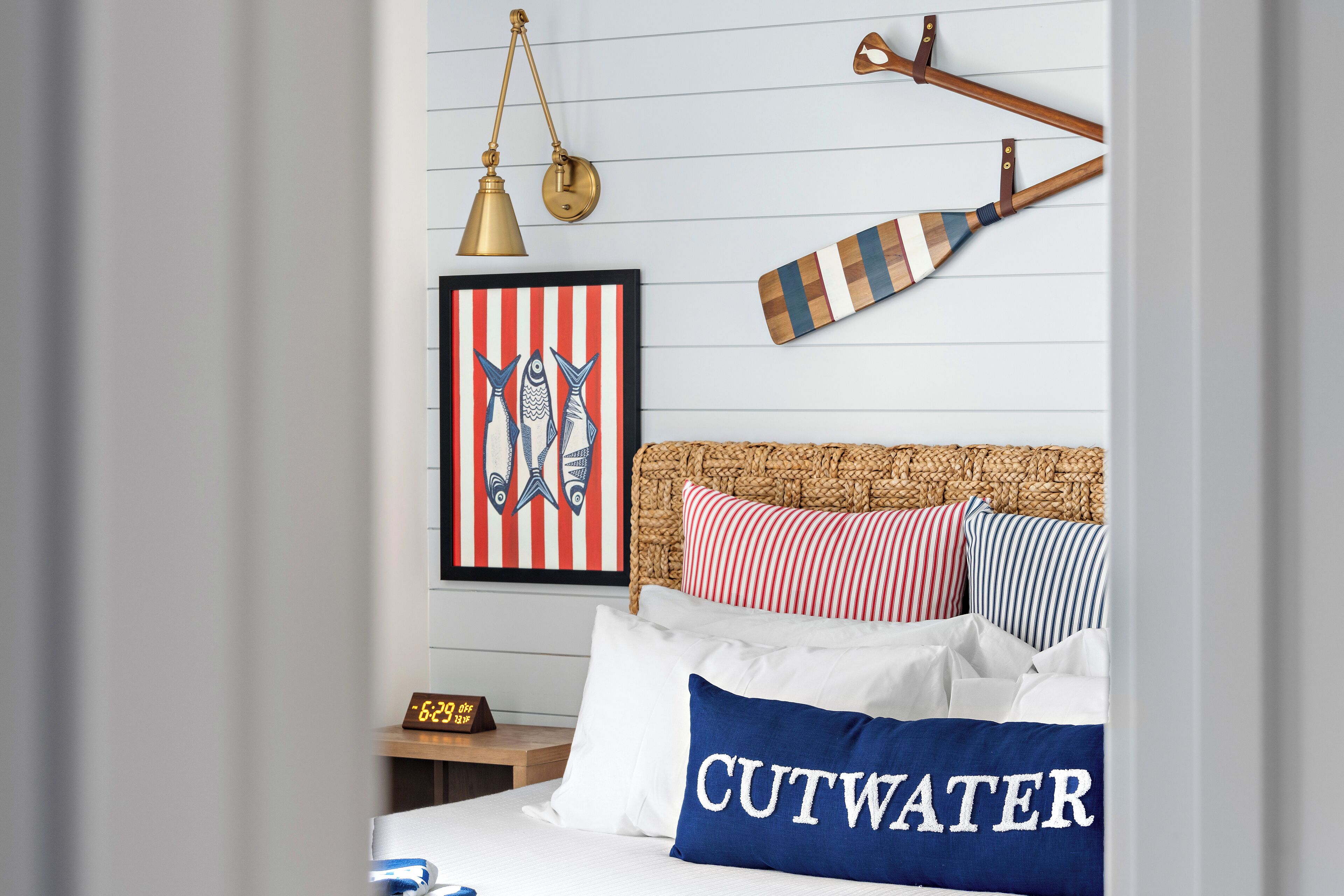 A neatly made bed with a pillow labeled "CUTWATER," a framed picture of fish, a wooden paddle, and a brass lamp.
