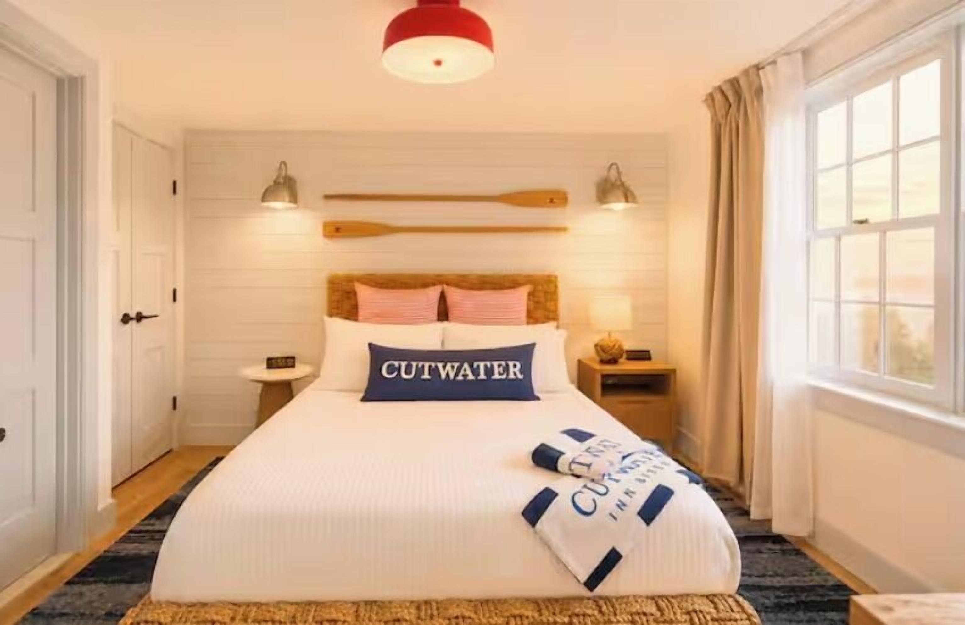 A hotel room with a bed, a bedside table, a nightstand, a window with curtains, and a door.