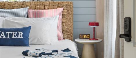 Una cama bien tendida con un cojín azul y blanco a rayas, un cabecero tejido y una mesita de noche con un reloj.