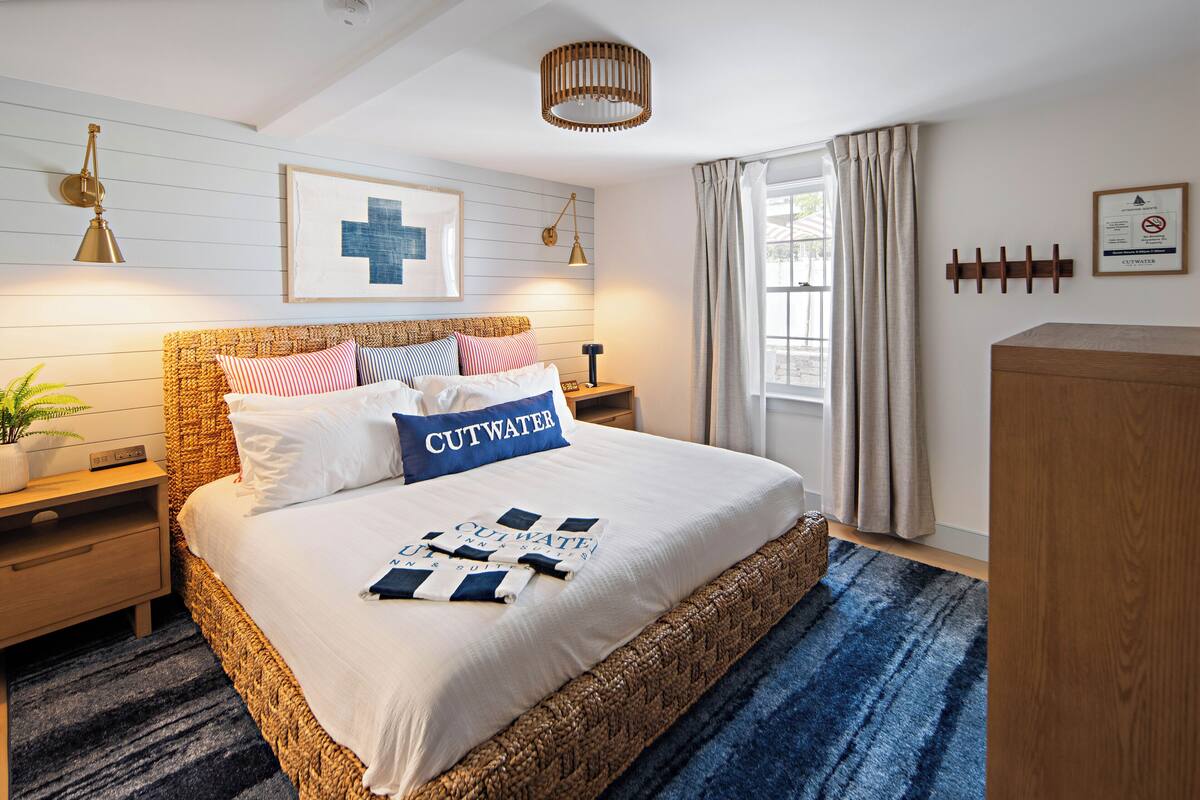 A neatly arranged bedroom with a large bed, a bedside table, a nightstand, a wall-mounted lamp, a framed picture, a window with curtains, and a wooden cabinet.