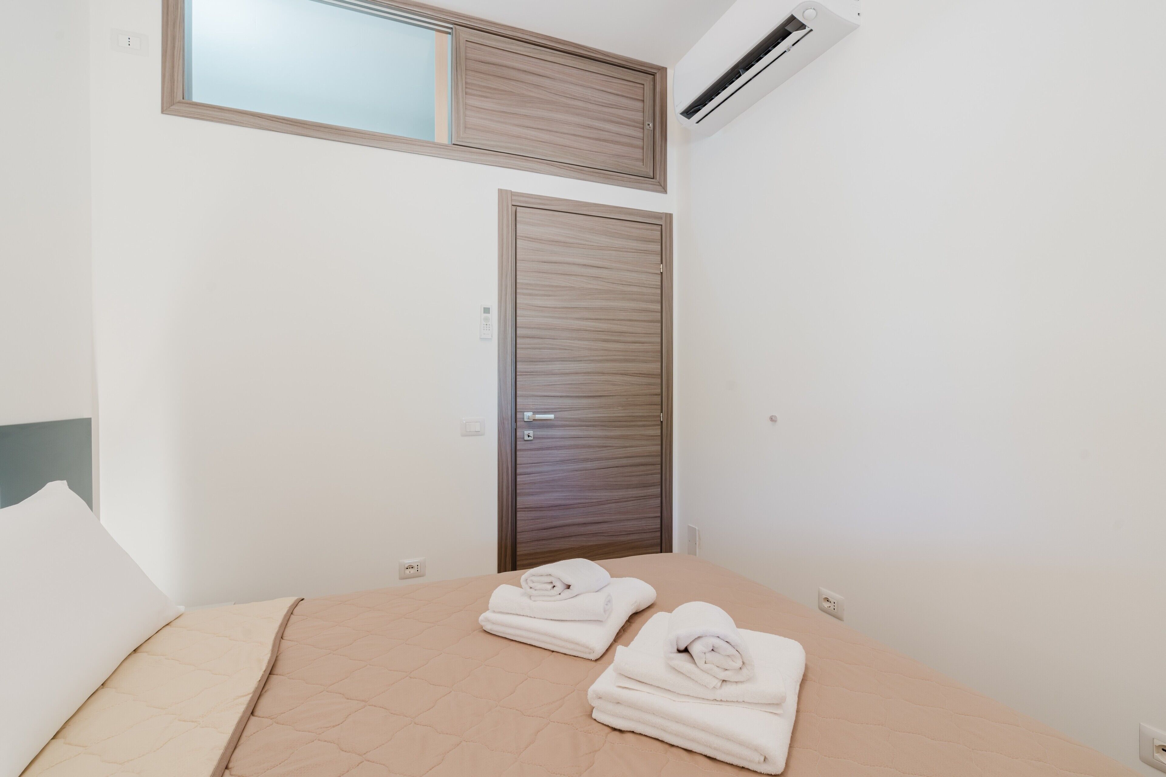 Un dormitorio con una cama, dos toallas, una ventana, aire acondicionado y una puerta de madera.