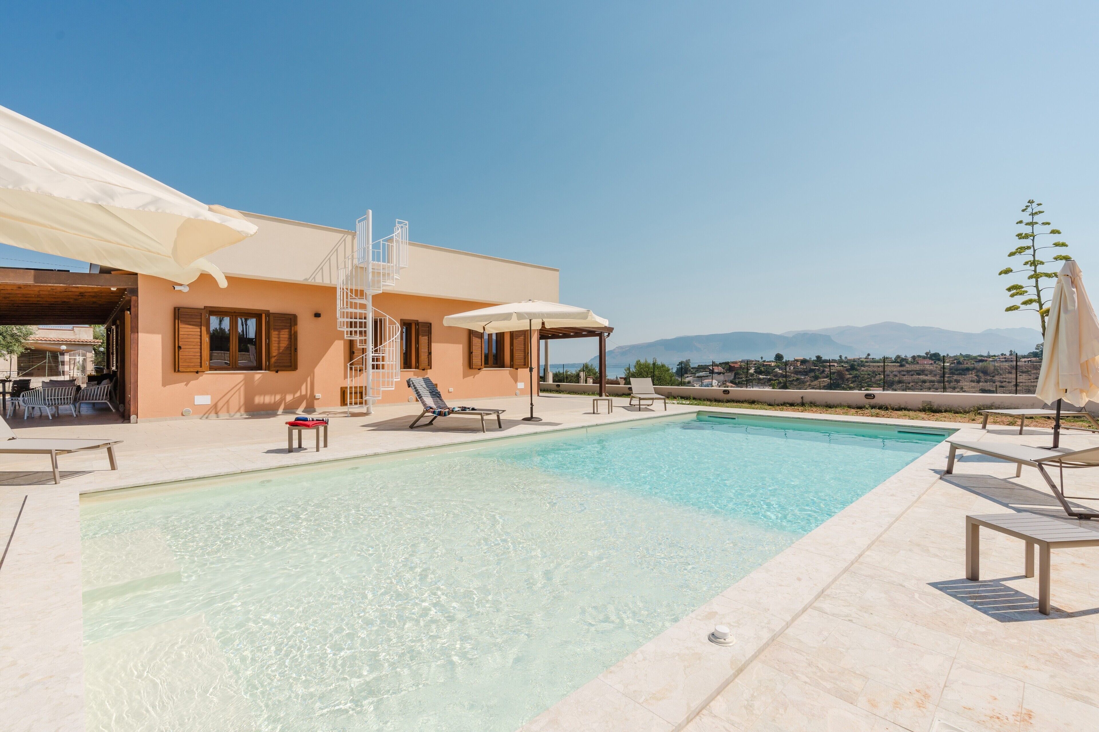 A pool area with lounge chairs, umbrellas, and a building with a staircase.