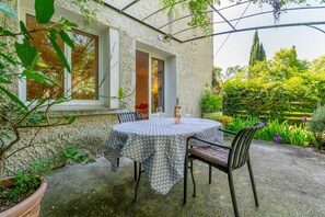A covered patio with a round table and chairs, a potted plant, and a window overlooking a garden.