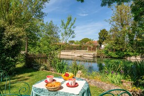 A table set with a basket, a bowl of fruit, and cups, overlooking a calm water body with greenery and a wooden fence in the background.