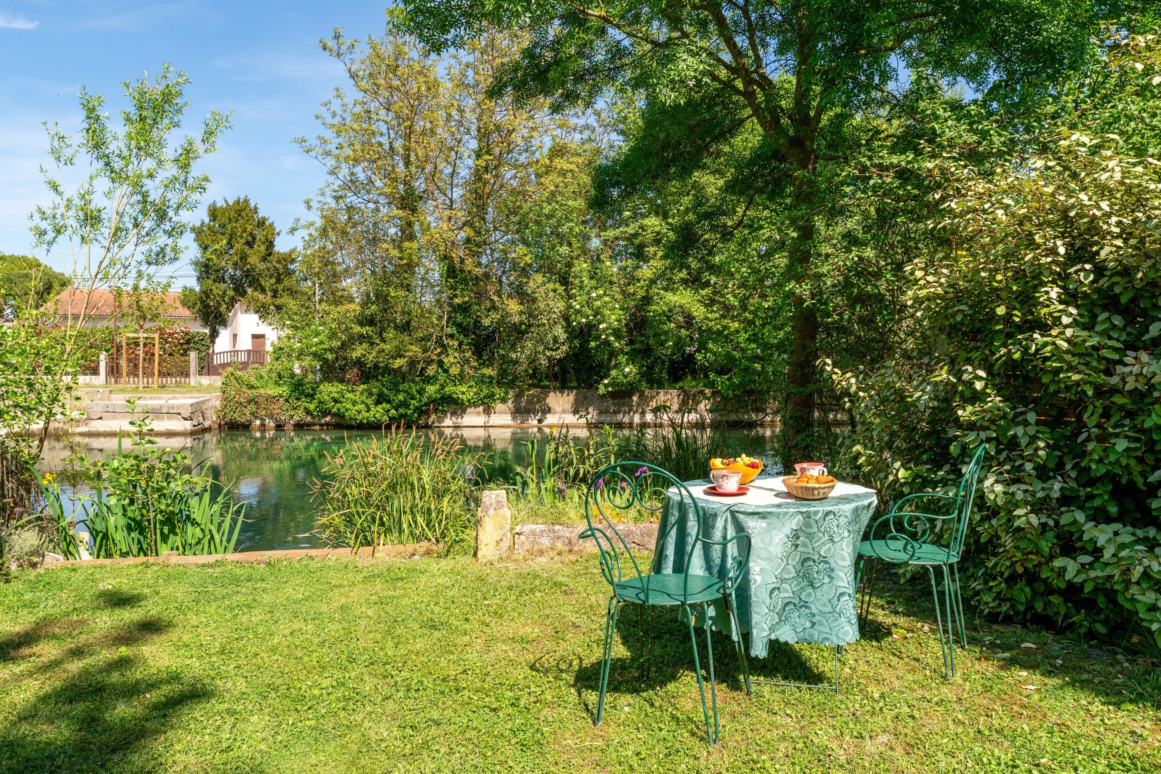 A serene outdoor dining area with a table and chairs, a pond, and lush greenery.