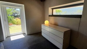 Et værelse med et hvidt kommode, et vindue med udsigt til grøn vegetation, og en dør der fører udendørs.