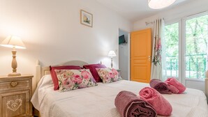 Un lit bien fait, agrémenté de coussins à motifs floraux, une table de chevet en bois avec une lampe, et une fenêtre avec des rideaux.
