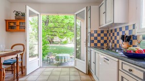 Une cuisine avec une porte ouverte donnant sur un jardin, une table à manger avec des chaises et un panier de fruits posé sur le comptoir.