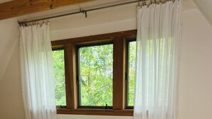 Un dormitorio con una cama grande, ropa de cama blanca, una mesita de noche de madera, un jarrón rústico y una ventana con vistas a árboles.