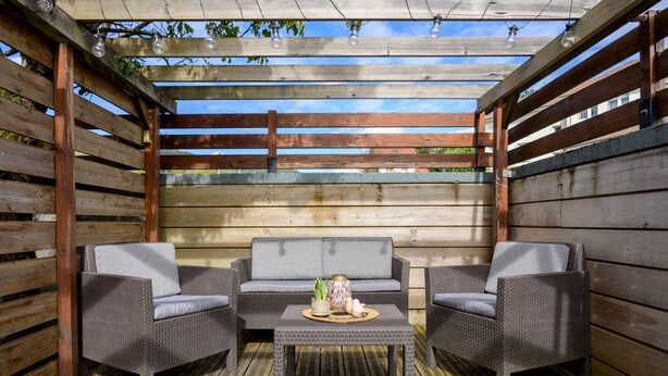 Ein Sitzbereich im Freien mit Korbmöbeln und einem Tisch, unter einer Holz-Pergola mit Lichterketten.