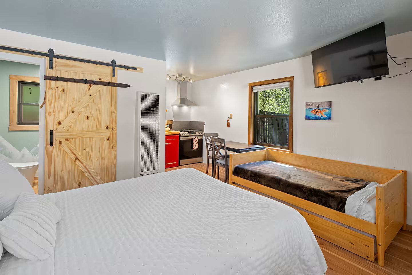 Una habitación compacta con una cama, una kitchenette pequeña, una puerta de madera y un televisor montado en la pared.