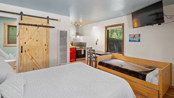 Una habitación compacta con una cama, una kitchenette pequeña, una puerta de madera y un televisor montado en la pared.