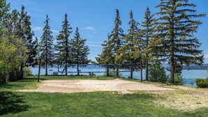 A grassy area with picnic tables and a volleyball net, surrounded by tall trees and a lake.