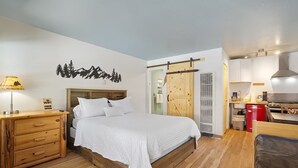 A bedroom with a wooden bed, a nightstand, a lamp, a wall-mounted artwork, a wardrobe, and a kitchenette with a stove.