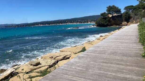 A coastal walkway with a wooden deck, rocky shoreline, and a view of the sea and distant buildings. - Top: Detached Villa for 8 People • Air Conditioning • Terrace • Beaches within a 10-minute walk (Saint-Cyr-sur-Mer)