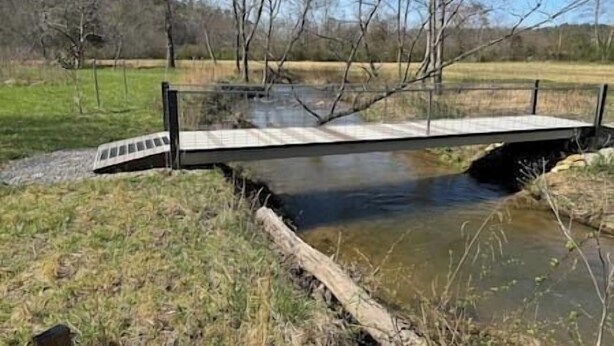 Un pont sobre un riu tranquil amb vores de gespa i arbres al fons.