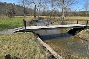 A bridge over a calm river with grassy banks and trees in the background. - Creekside Cabin Near Blue Ridge | Fire Pit +Nature (Mineral Bluff)