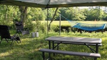 Montaje para picnic al aire libre con mesa y sillas bajo un toldo, una hamaca y un espacio cubierto de césped con árboles.
