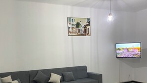 A living room with a grey sofa, a small white table, a TV on the wall, and a picture of a street scene. - House with private pool close to town center (Houmet souk)