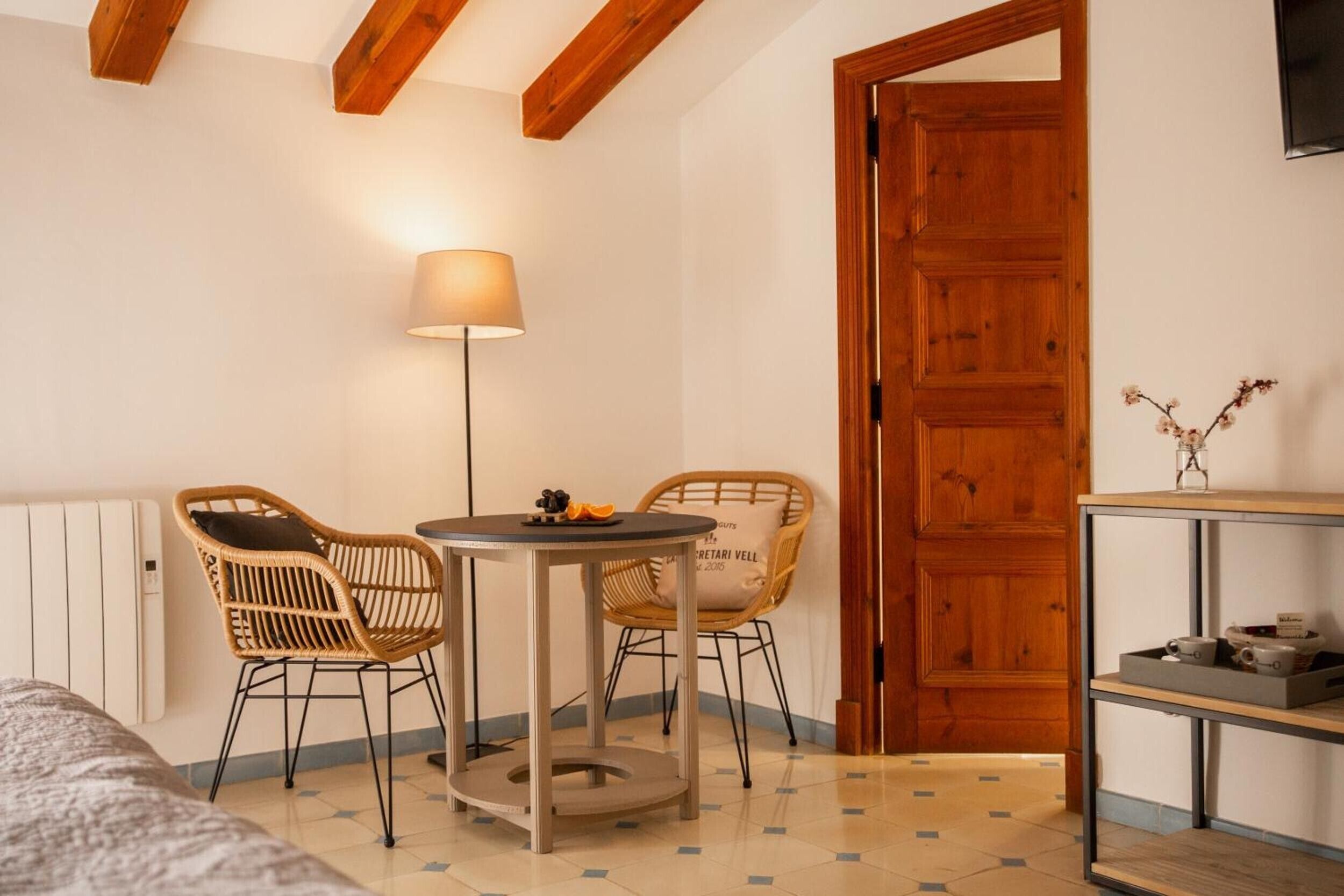 A room with a round table, two wicker chairs, a floor lamp, a wooden door, and a shelf with items.