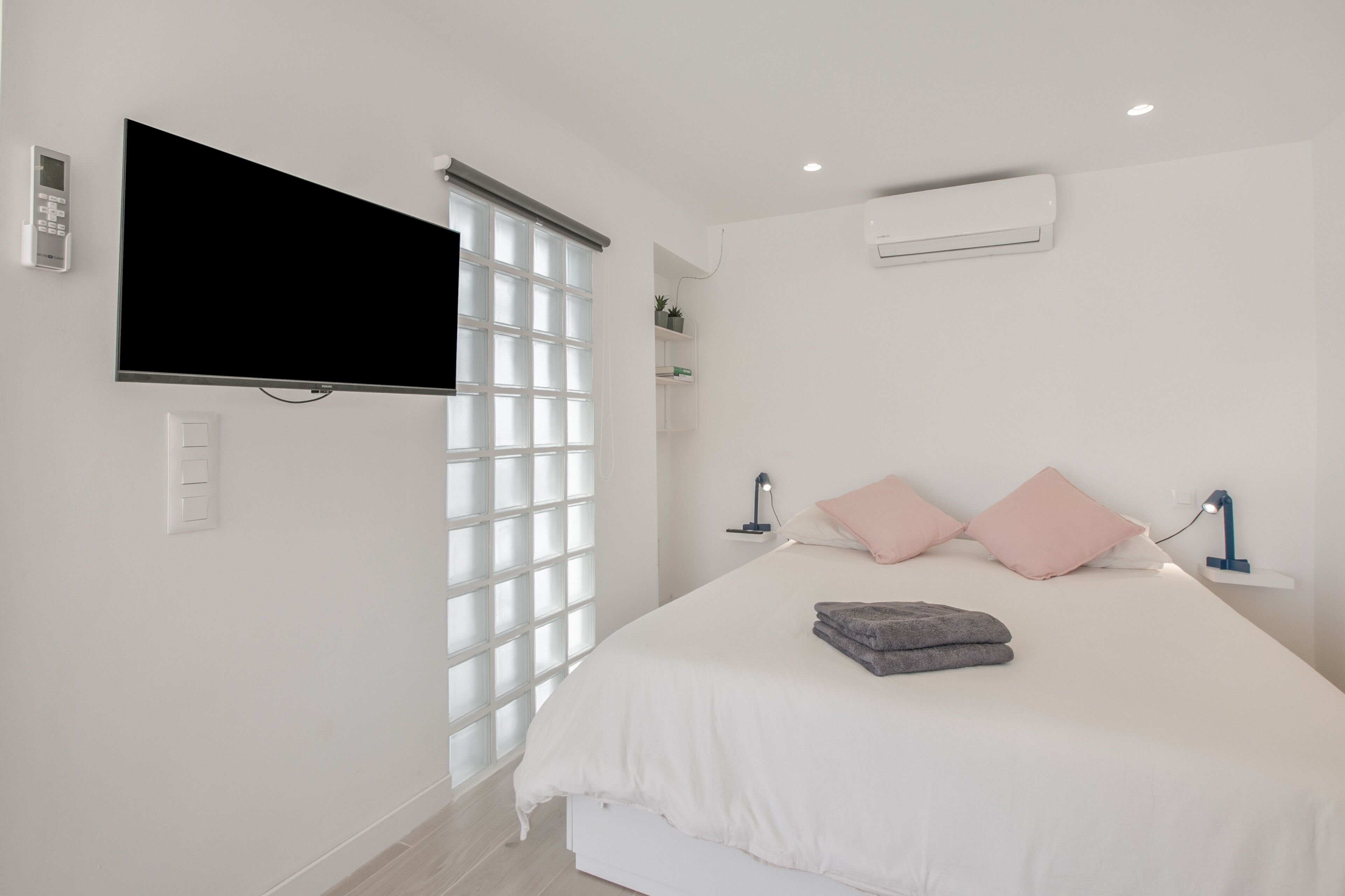 Un dormitorio con cama, televisión montada en la pared, aire acondicionado y ventana con persianas.