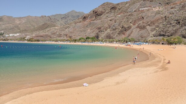 Ein Strand mit klarem Wasser, sandigem Ufer und Bergen im Hintergrund.