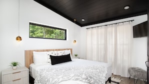 A modern bedroom with a large bed, a nightstand, a small table with a plant, a window with a view of greenery, and a ceiling with recessed lighting. - The Little Timber: Cozy, Hot Tub & Fireplace! (Townsend)