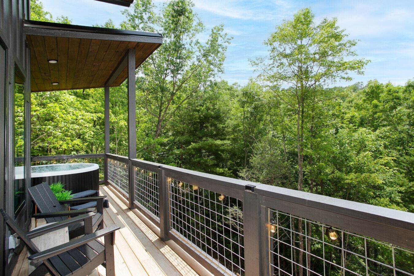 A wooden deck with a table and chairs, surrounded by a metal railing, overlooking a forested area.