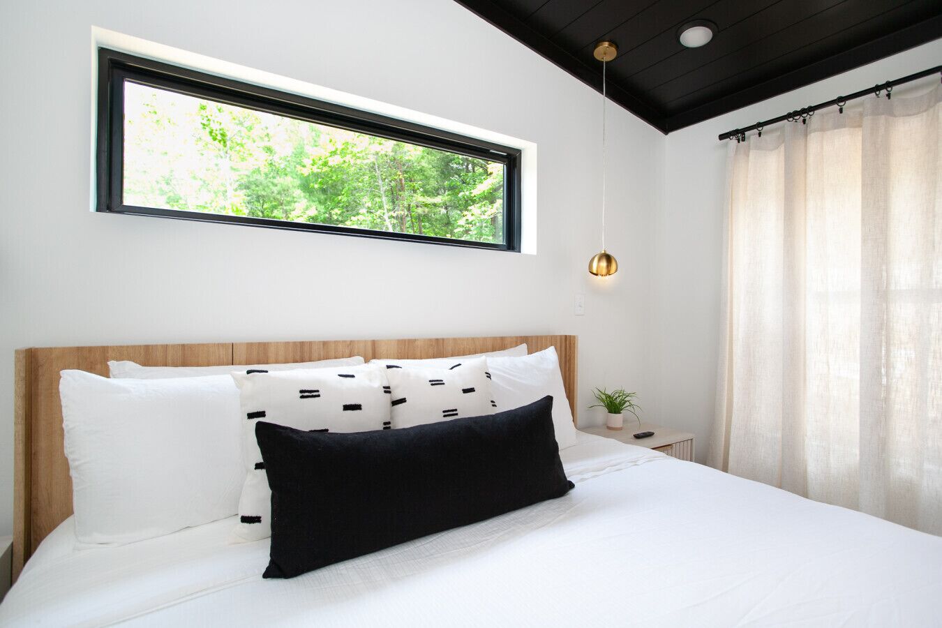 Une chambre avec une grande fenêtre, un lit avec une literie blanche et noire, une tête de lit en bois, une petite table de chevet avec une plante et un luminaire suspendu.