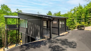 A cabin with a covered porch and a small attached structure, surrounded by dense forest. - The Little Timber: Cozy, Hot Tub & Fireplace! (Townsend)