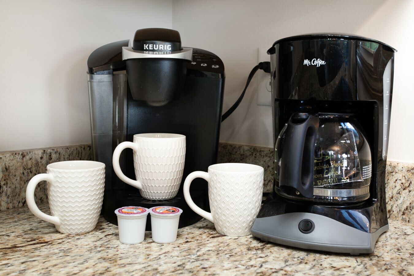 A Keurig coffee maker with two white mugs and two small containers of coffee pods on a granite countertop.