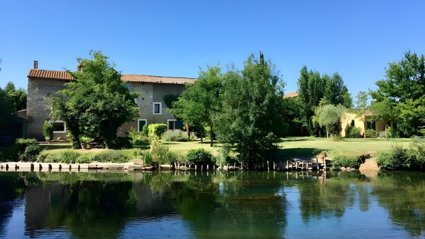 A serene lakeside setting with a stone building, lush greenery, and a clear reflection in the water.