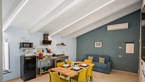 A compact kitchen with a microwave, stove, and sink, a dining table with yellow chairs, and a blue sofa in a room with light blue walls and wooden flooring.