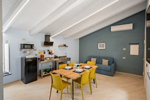 A compact kitchen with a microwave, stove, and sink, a dining table with yellow chairs, and a blue sofa in a room with light blue walls and wooden flooring. - Apartment 'Deluxe Gigli' with Wi-Fi and Air Conditioning (Ascea)