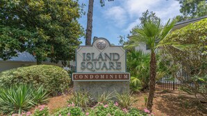 A sign for Island Square Condominium surrounded by lush greenery and flowering plants. - Centrally located condo for 4 with a community swimming pool. (Saint Simons Island)