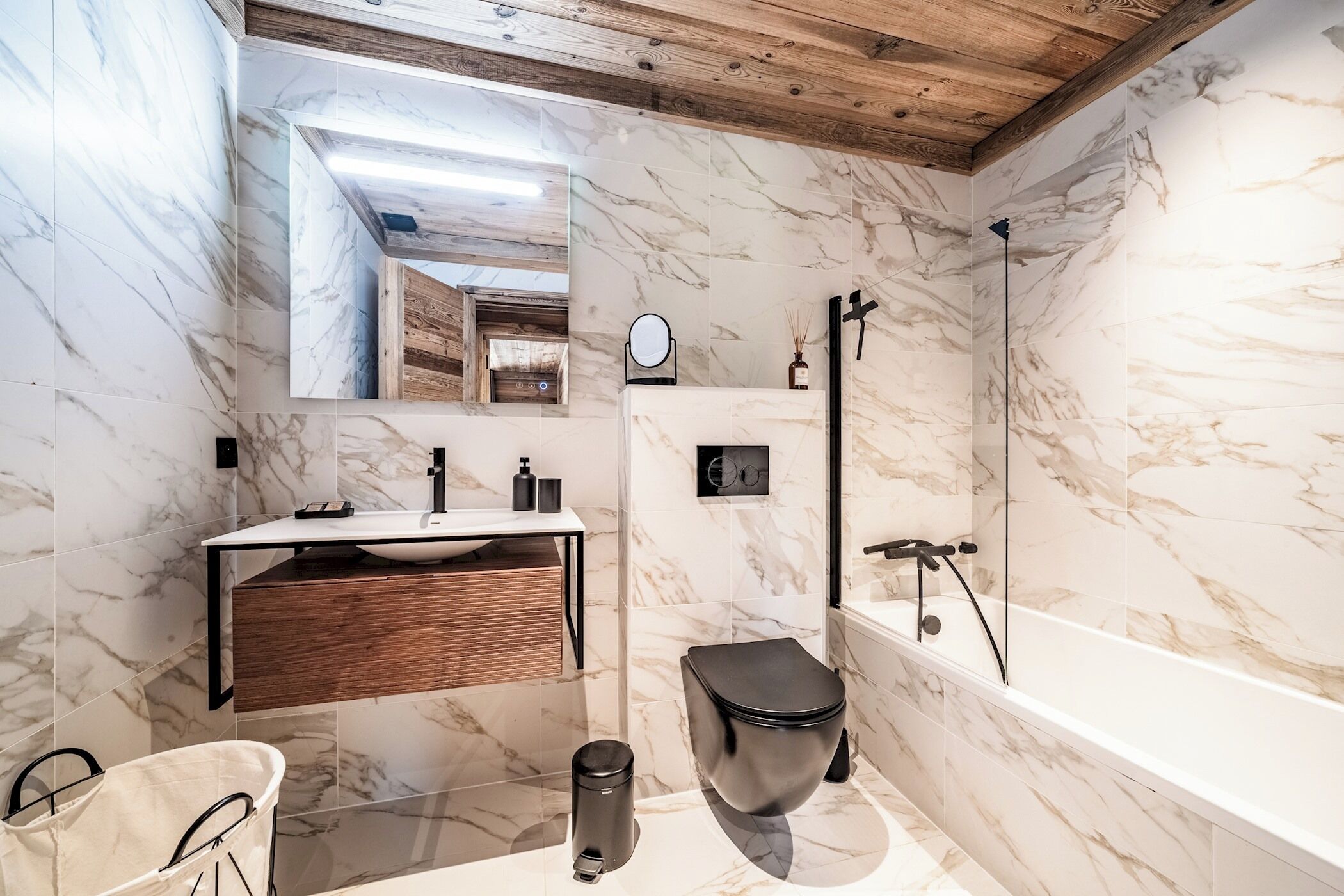 Ein modernes Badezimmer mit Marmorwand, Holzboden, einer schwarzen Toilette, einer Badewanne und einem Glaswaschbecken.
