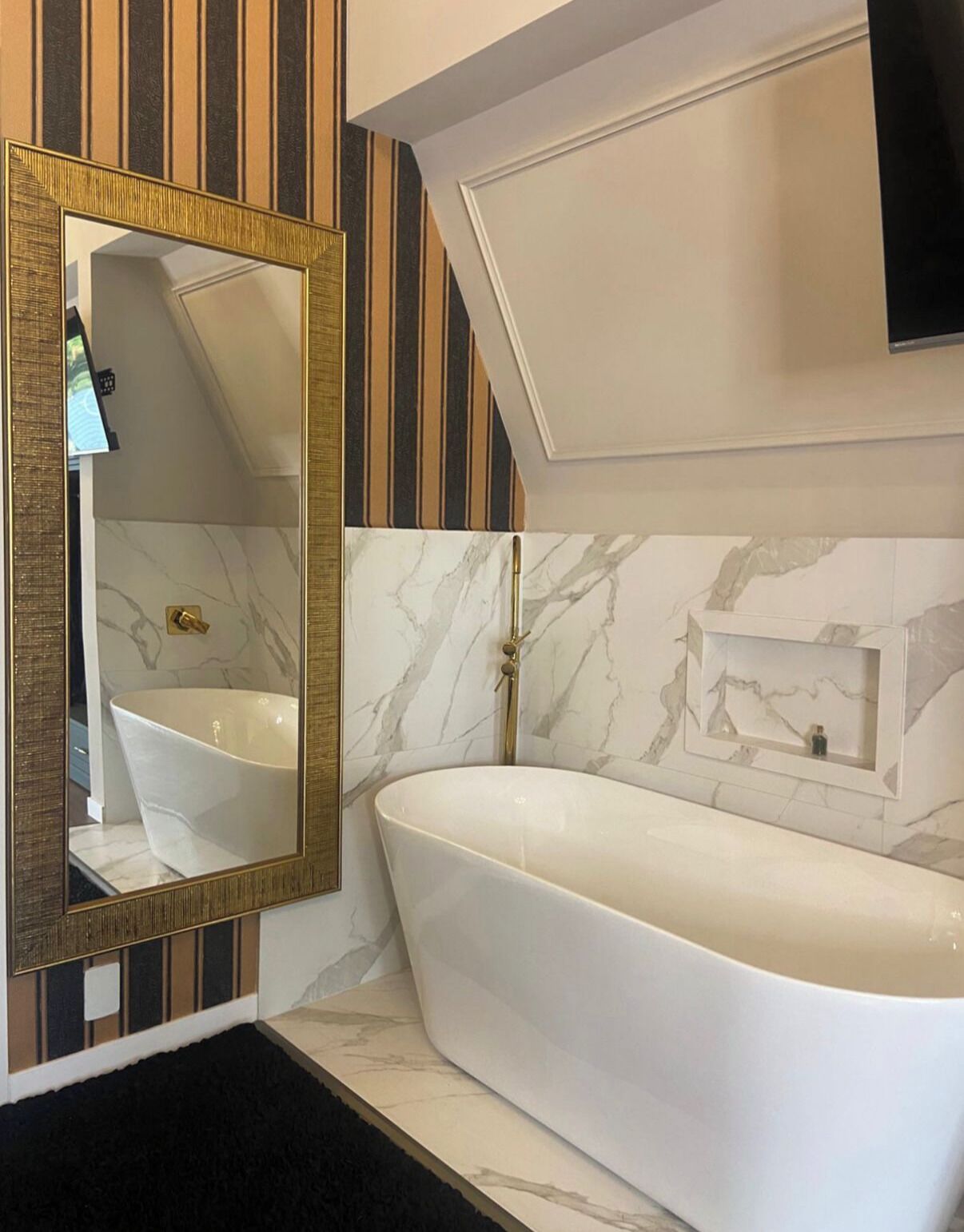Un baño moderno con una bañera independiente, un espejo grande con marco dorado y paredes con diseño de mármol.
