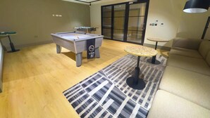 Una mesa de billar, una mesa de fútbol de mesa, una mesita y un sofá en una habitación con suelo de madera y una alfombra estampada.