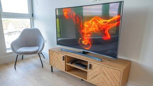 Una sala de estar moderna con un televisor de pantalla plana sobre un mueble de madera, un sillón gris y un ventanal grande.