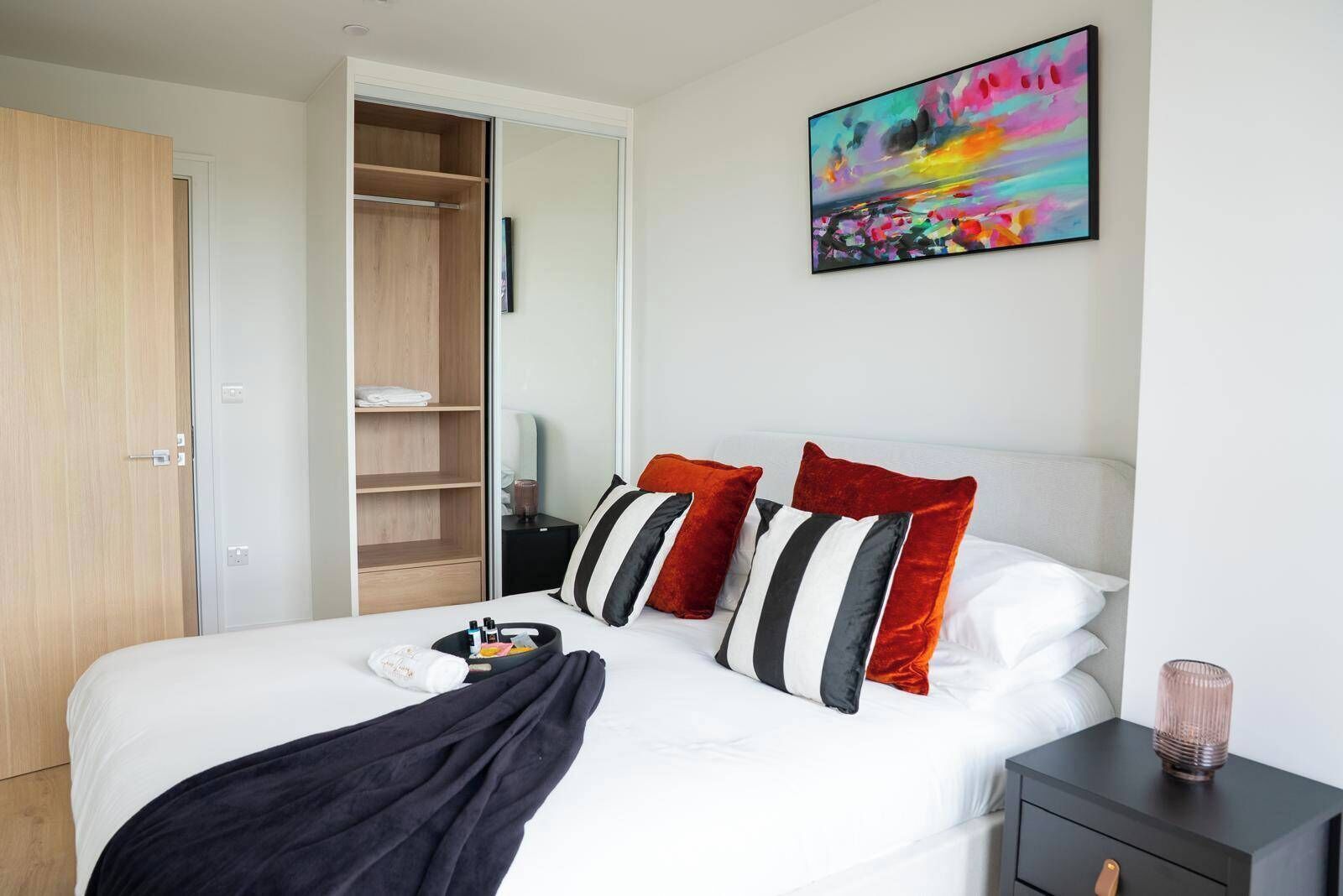 A hotel room with a bed, a bedside table, a nightstand, a wardrobe, and a colorful abstract painting on the wall.