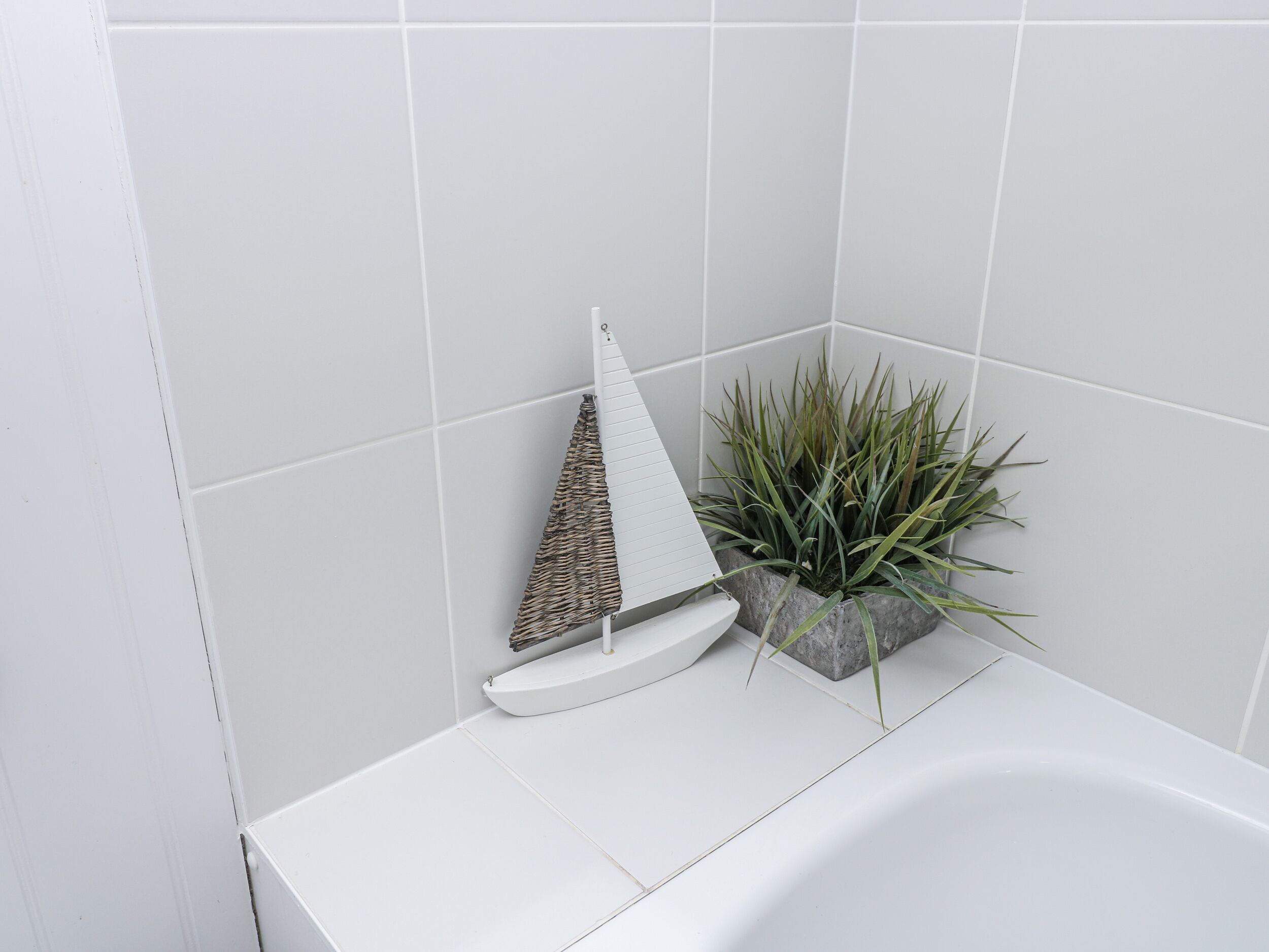 Ein Badezimmer mit weißen Fliesen, einer weißen Badewanne, einem dekorativen Segelboot und einem Blumentopf.