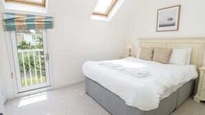 Une chambre à coucher comprenant un lit, une table de chevet, une lampe, un tableau encadré, une fenêtre donnant sur un paysage verdoyant et une porte menant à un balcon.
