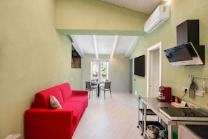 Una zona de estar amb un sofà vermell, taula de menjador amb cadires, zona de cuina amb fogó i un televisor fixat a la paret.
