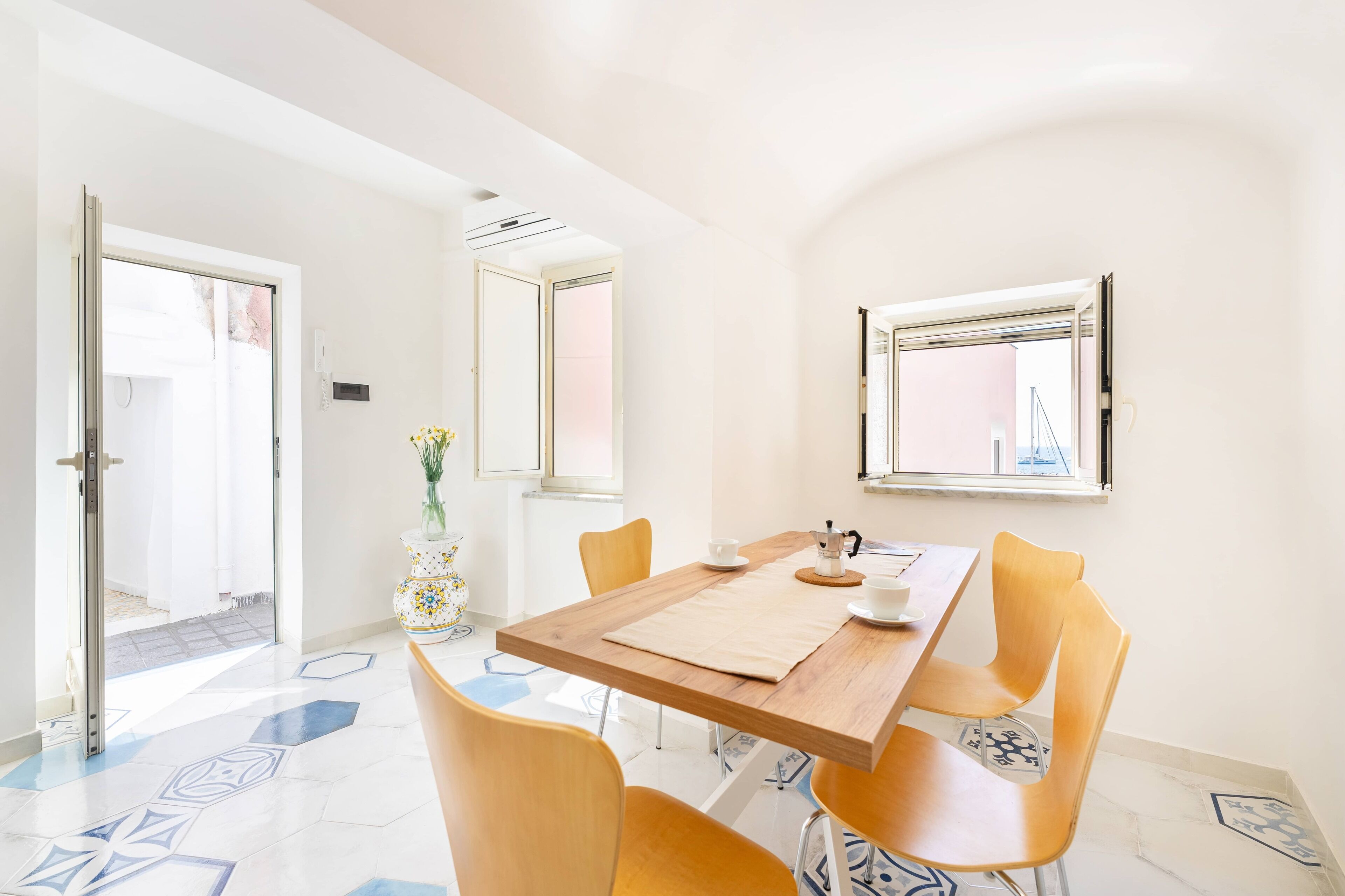 Un comedor con una mesa de madera, cuatro sillas amarillas, un jarrón con flores y una ventana que ofrece vistas a un puente.