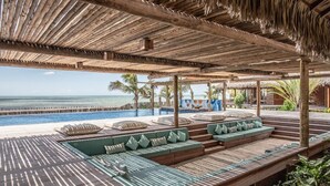 A poolside area with wooden deck seating, a thatched roof, and a view of the ocean. - Kabana Hotel (Cruz)